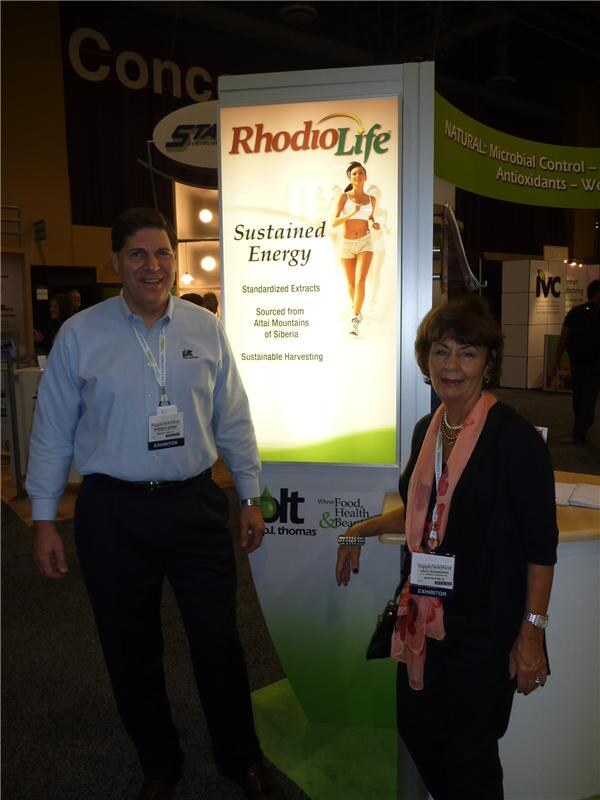 A man in a light blue shirt and a woman in black with a coral scarf stand beside a lit RhodioLife sign reading Sustained Energy at the P.L. Thomas booth.