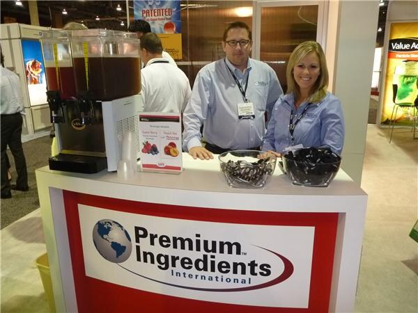 A man with glasses and a smiling woman in blue shirts stand behind a Premium Ingredients International counter with beverage dispensers and bowls of dark snacks.