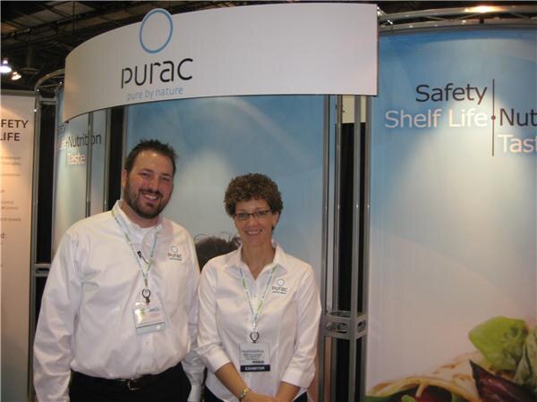 A bearded man and a woman with glasses, both in white shirts with Purac logos, smile in front of the Purac booth with a pure by nature sign.