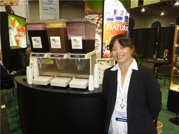 A smiling woman in a black blazer with an exhibitor badge stands beside three RFI beverage dispensers filled with dark drinks at the booth.
