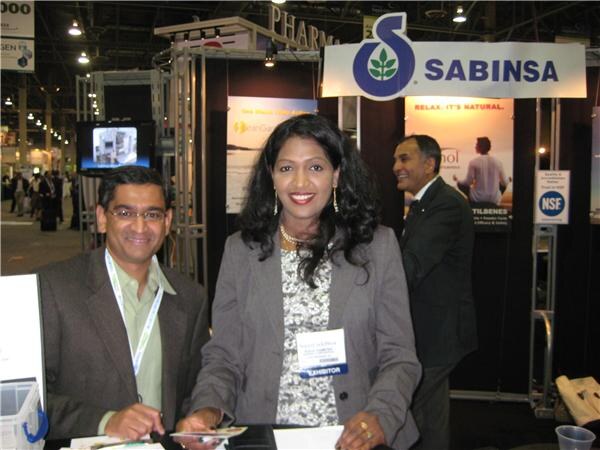 A man with glasses and a smiling woman in a gray blazer pose in front of the Sabinsa sign, with another man standing in the background.