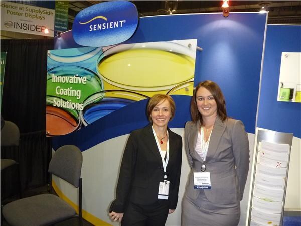 Two women in dark business jackets with exhibitor badges stand in front of a Sensient booth wall reading Innovative Coating Solutions, with colorful paint-like images.