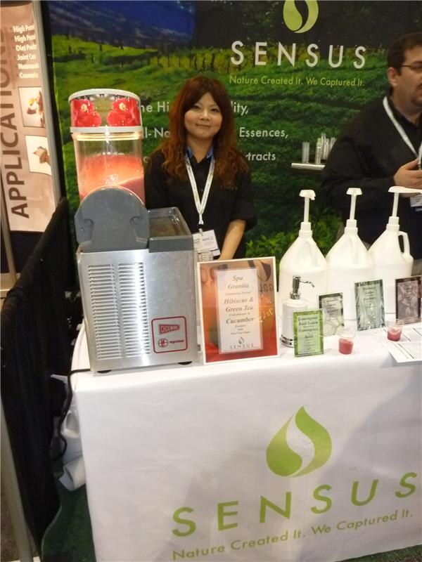 A woman in a black top with a lanyard stands behind a Sensus table beside a slush machine with red drink, with a man partly visible at right and bottles and signs on the table.