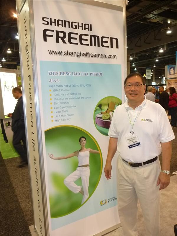 A smiling man in a white polo and exhibitor badge stands beside a tall Shanghai Freemen banner about high purity stevia Reb-A, with other attendees in the background.