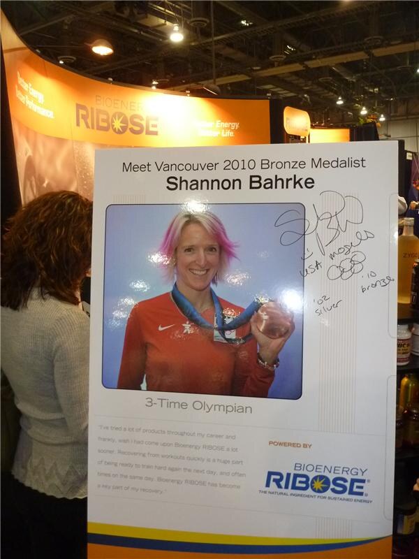 A poster at the Bioenergy Ribose booth reads Meet Vancouver 2010 Bronze Medalist Shannon Bahrke, showing her holding a medal, with a signature. A woman with curly hair stands at left.