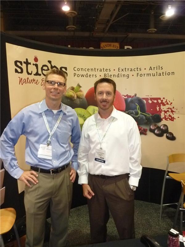 Two men in dress shirts with badges and lanyards stand in front of a curved Stiebs backdrop listing concentrates, extracts, arils, powders, blending and formulation, with fruit images.