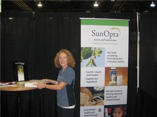 A smiling woman with curly hair leans on a small table with a Food Product Design display, beside a SunOpta Grains and Foods Group banner about soy foods, in a booth with black curtains.