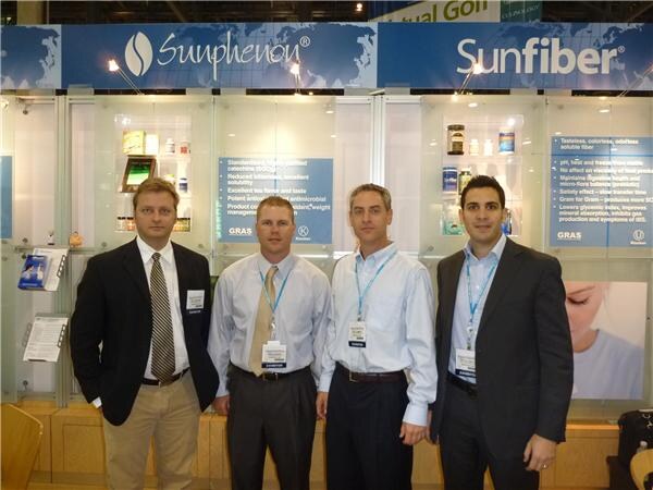 Four men in business attire with badges stand in front of a booth with Sunphenon and Sunfiber signs, product displays and information panels.