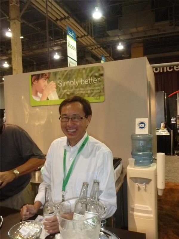 A smiling man in glasses and a white shirt with a green lanyard sits at a table with bottles in ice, below a Sweet Green Fields sign reading Simply better; a water cooler is at right.