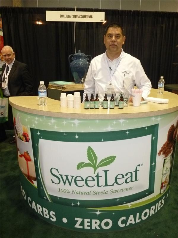 A man in a white shirt stands behind a curved SweetLeaf counter with small dropper bottles and cups; the front reads 100% Natural Stevia Sweetener, Zero Carbs, Zero Calories.