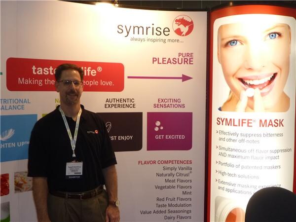 A man with glasses in a black polo and exhibitor badge stands in front of Symrise signage, including a Symlife Mask panel with a smiling woman's face.