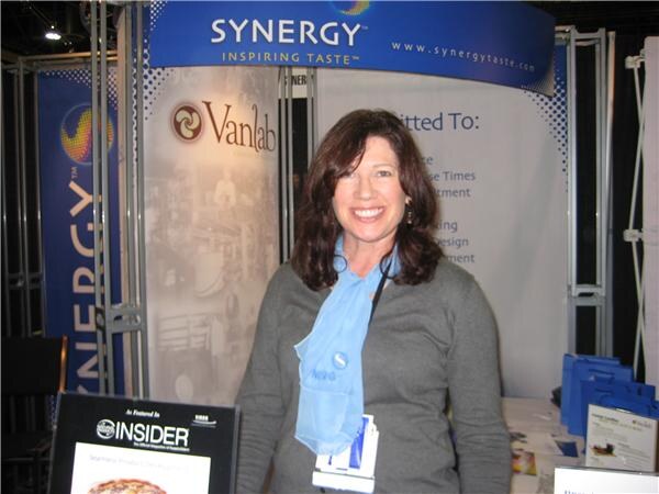 A smiling woman with long brown hair and a blue scarf stands in front of Synergy and Vanlab signs, with an Insider display on a table in front of her.