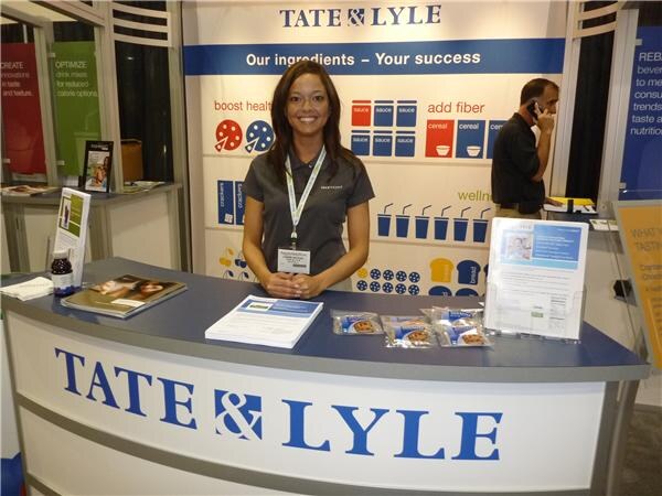 A smiling woman in a dark polo and lanyard stands behind the curved Tate & Lyle booth counter, with brochures and snack packets on it and a banner reading Our ingredients – Your success behind her.