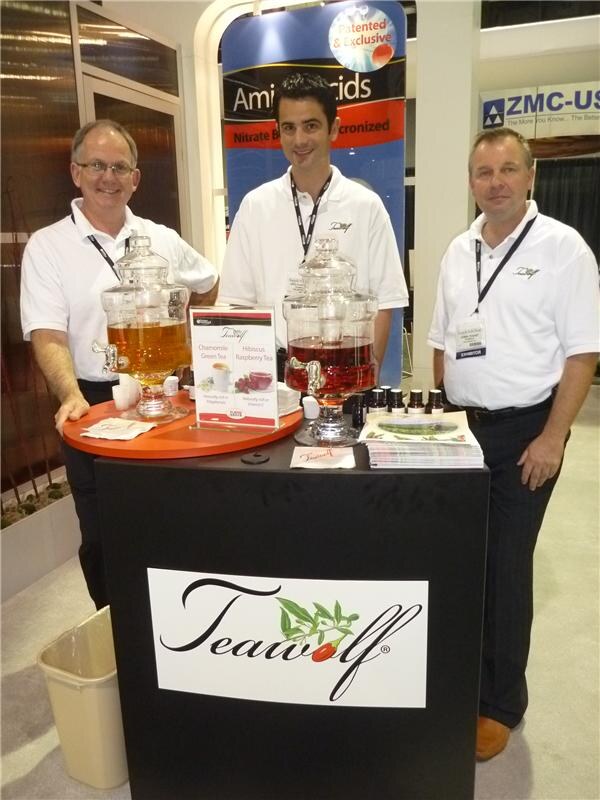 Three men in white polo shirts and lanyards stand around a Teawolf display with two glass dispensers of iced tea, a menu card and bottles; a ZMC-USA sign is visible behind them.