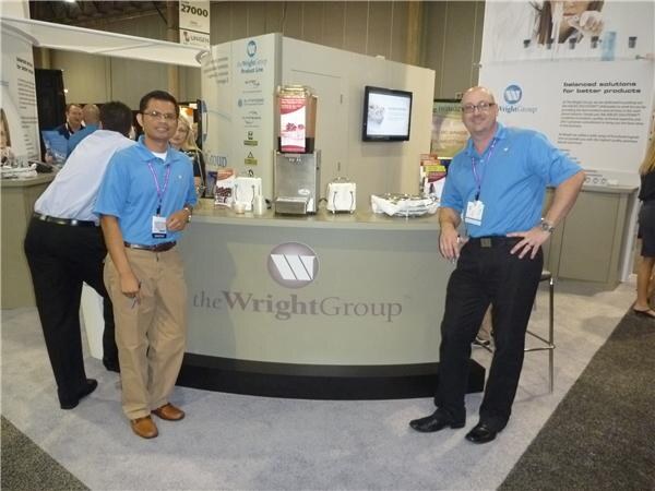 Two men in blue polo shirts and lanyards stand beside the curved The Wright Group booth counter, which holds a beverage machine, small appliances and a screen; other attendees stand behind.