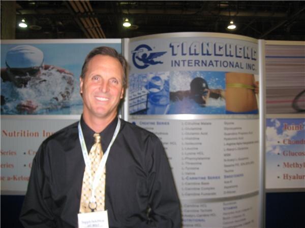 A smiling man in a dark shirt, patterned tie and lanyard stands in front of Tiancheng International Inc. display panels listing ingredient series, with swimming photos on the panels.