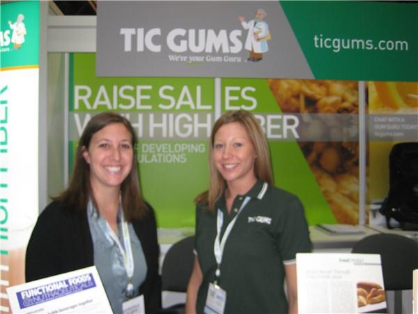 Two smiling women wearing lanyards stand in front of a green Tic Gums banner; one wears a dark Tic Gums polo, and trade magazines sit in the foreground.