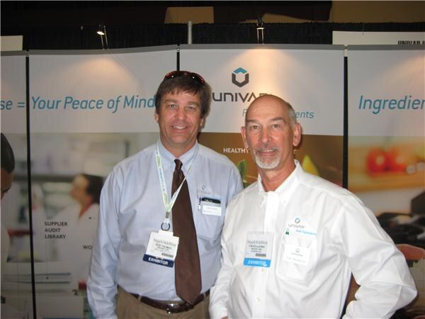 Two men wearing exhibitor badges stand side by side in front of Univar banners; one in a light blue shirt and tie, the other in a white shirt with a gray goatee.