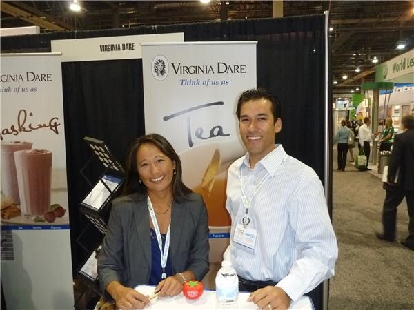 A smiling woman in a gray blazer and a man in a striped shirt, both with lanyards, stand at a table in front of Virginia Dare banners, one reading Think of us as Tea.