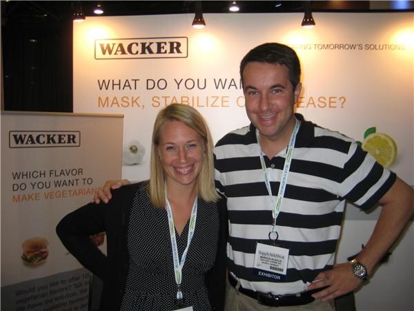 A smiling woman in a polka-dot top and a man in a striped shirt with an exhibitor badge pose together in front of Wacker signs, one about making flavors vegetarian.