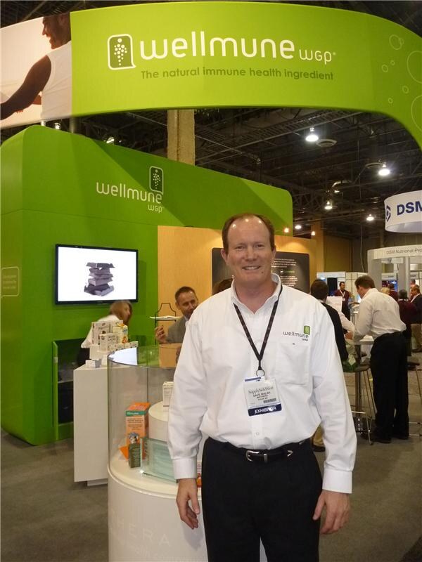 A man in a white Wellmune shirt with an exhibitor badge stands in front of the green Wellmune WGP booth, which has a screen and product displays; other people talk in the background.