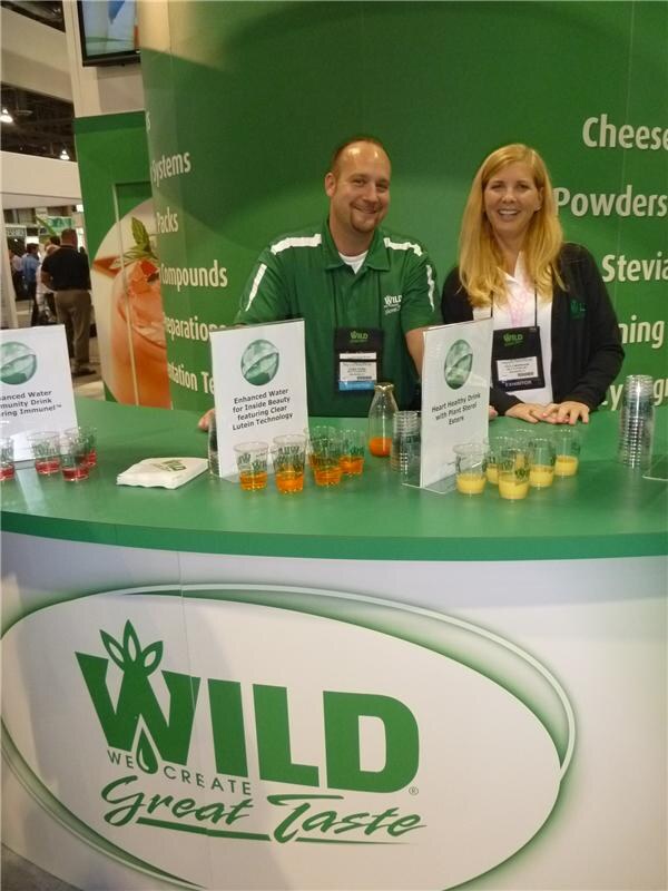 A man in a green polo and a woman with long blond hair stand behind a green WILD counter with sample cups of orange and yellow drinks and signs about enhanced water and a heart healthy drink.