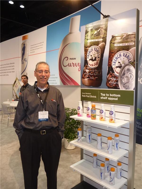 A man in a dark gray shirt with a lanyard stands beside an Avery Dennison display of labeled bottles on shelves. Behind him are Fasson graphics and a poster of French Vanilla coffee cans.