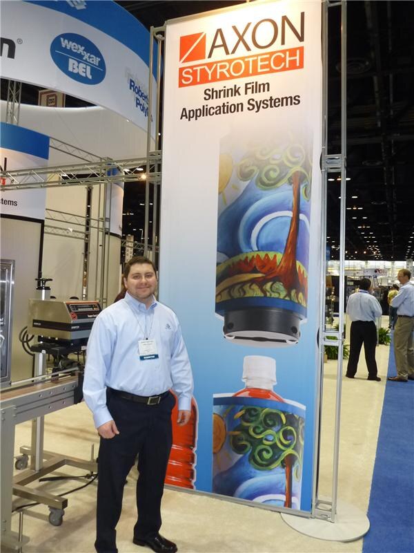 A smiling man in a light blue shirt stands in front of a tall Axon Styrotech banner reading Shrink Film Application Systems. A Wexxar Bel sign and a machine are visible behind him.