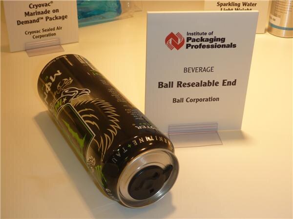 A black energy drink can with a resealable end lies on its side beside an Institute of Packaging Professionals sign for Ball Resealable End, with a Cryovac Sealed Air sign behind.