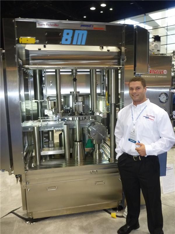 A man in a white shirt stands beside a large stainless steel machine marked Barry-Wehmiller and 8M. An Angelus machine is partly visible behind him on the right.