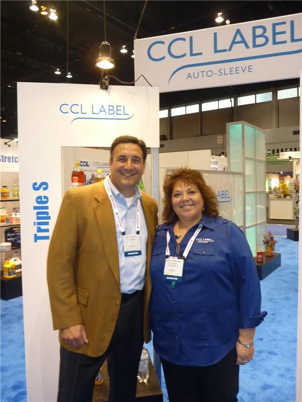 A man in a tan blazer and a woman in a blue CCL Label shirt stand together smiling at a CCL Label Auto-Sleeve booth. Bottles are displayed in an alcove behind them.