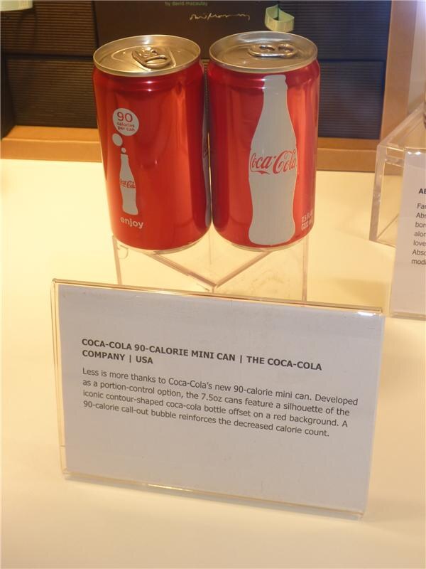 Two red 7.5oz Coca-Cola mini cans, one with a 90 calories per can bubble, sit on a clear stand above a card describing the Coca-Cola 90-calorie mini can.