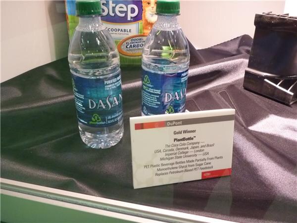 Two Dasani water bottles with PlantBottle logos on black fabric, in front of a DuPont Gold Winner sign for PlantBottle naming The Coca-Cola Company.