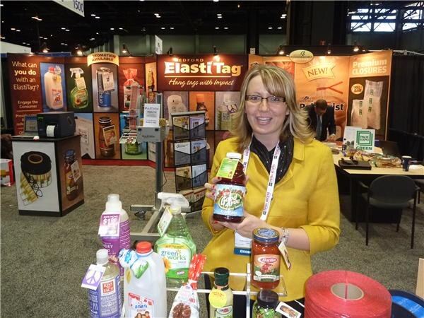 A smiling woman in a yellow jacket holds a Barilla sauce jar with a hang tag at the ElastiTag booth. Green Works and other tagged bottles are in front of her.