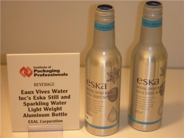 Two silver aluminum bottles labeled eska natural spring water stand beside a sign for Eaux Vives Water Inc's Eska light weight aluminum bottle by EXAL Corporation.