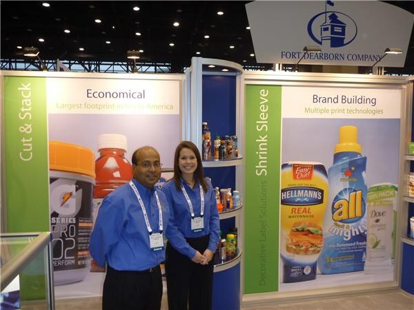A man and a woman in blue shirts stand at the Fort Dearborn Company booth. Panels behind them show labeled products including Hellmann's, all and Dove under Shrink Sleeve and Cut & Stack.