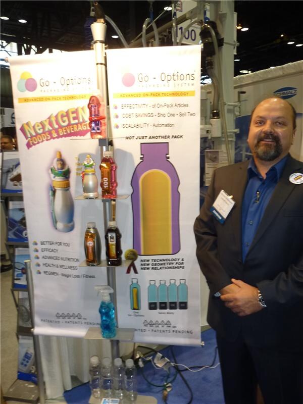 A bearded man in a dark blazer stands beside Go-Options banners about on-pack technology. Bottles with attached packaging are displayed on small shelves between the banners.