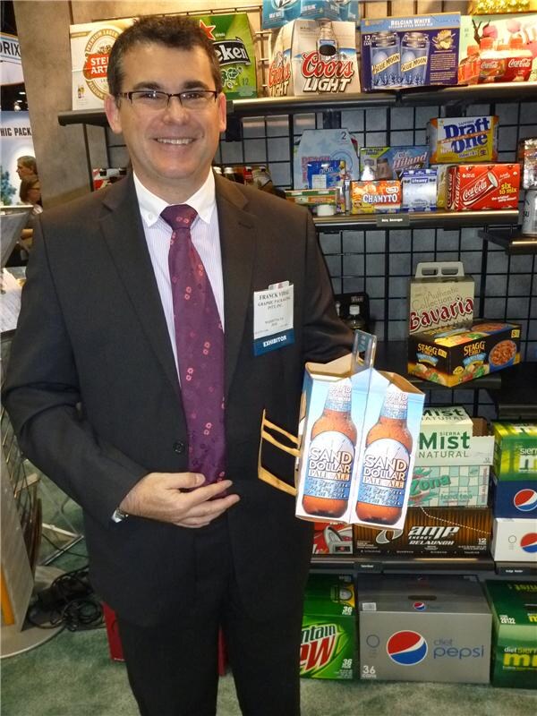 A smiling man in a dark suit and burgundy tie holds a Sand Dollar Pale Ale carrier. Shelves behind him hold cartons for Coors Light, Pepsi, Mountain Dew, Sierra Mist, AMP Energy, Stagg and Coca-Cola.