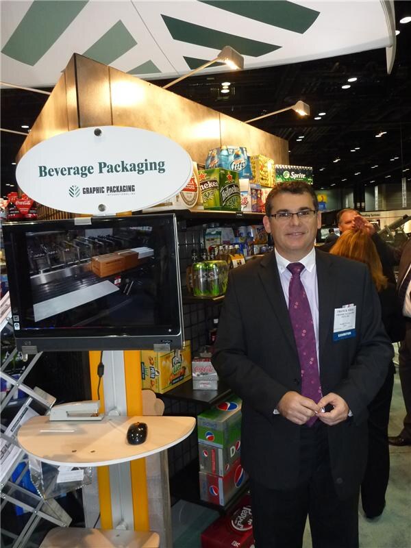 A man in a dark suit and burgundy tie stands beside a Graphic Packaging International Beverage Packaging sign and a monitor. Shelves behind hold Heineken, Sprite, Pepsi and Coca-Cola packs.