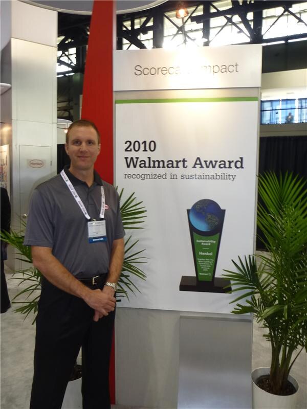 A man in a gray polo with a lanyard badge stands beside a sign reading 2010 Walmart Award recognized in sustainability, with a Henkel Walmart sustainability award trophy and potted palms.