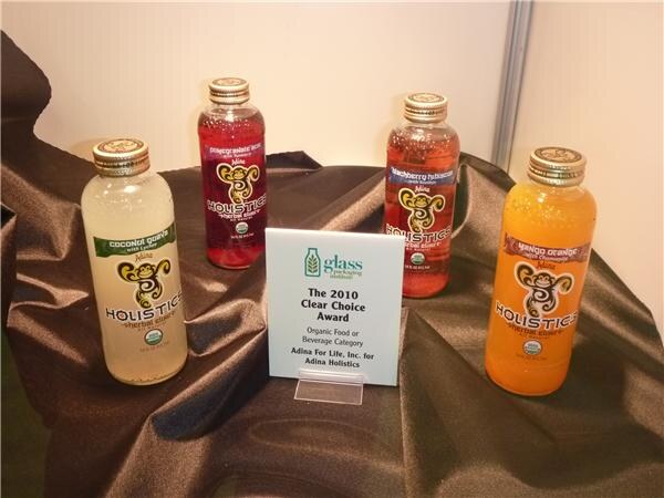 Four bottles of Holistics herbal elixir in coconut guava, pomegranate, blackberry hibiscus and mango orange flavors on brown fabric, beside a 2010 Clear Choice Award sign.