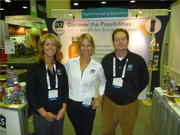 Two women and a man in dark and white shirts with ILS logos and exhibitor badges stand in front of an ILS booth sign reading Digital Packaging Specialists and Discover the Possibilities.
