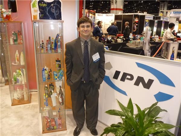 A young man in a gray suit and tie with a name badge stands beside the IPN booth counter, next to a tall glass display case of bottles and containers.