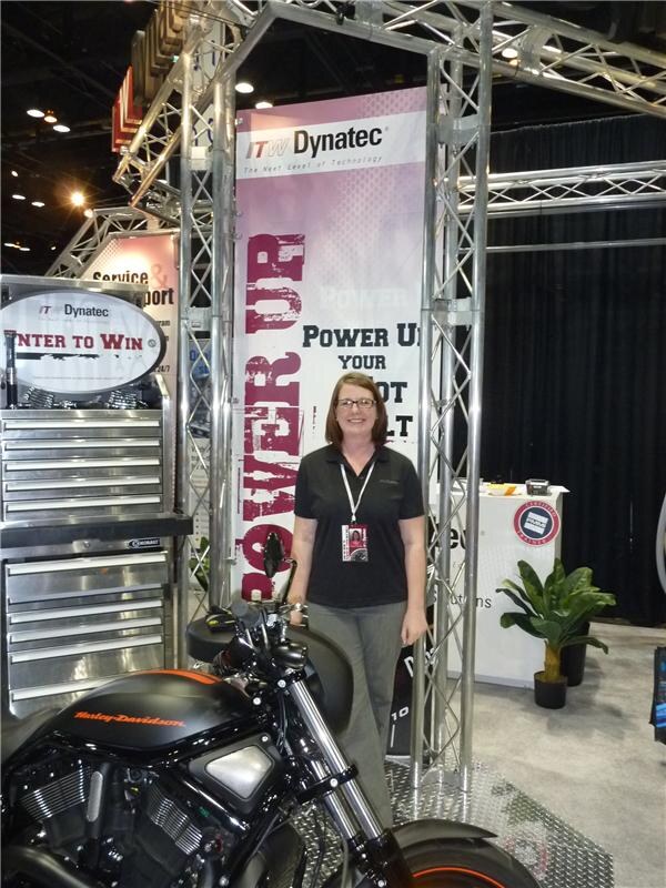 A woman in a black shirt and glasses stands under a truss at the ITW Dynatec booth. A Harley-Davidson motorcycle and a tool chest with an Enter to Win sign are in the foreground.