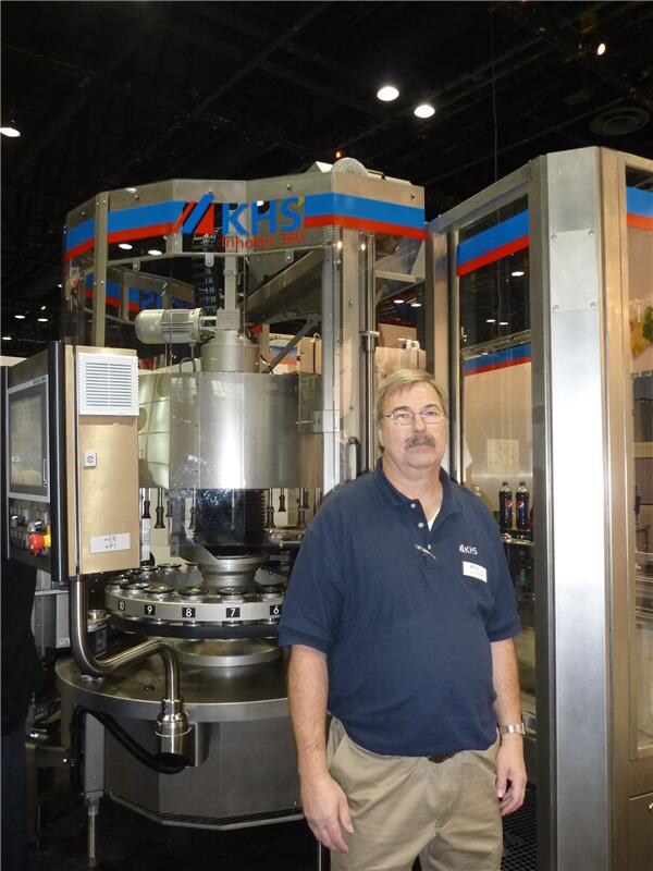 A mustached man with glasses, wearing a navy KHS polo and khaki pants, stands in front of a large KHS rotary filling machine.