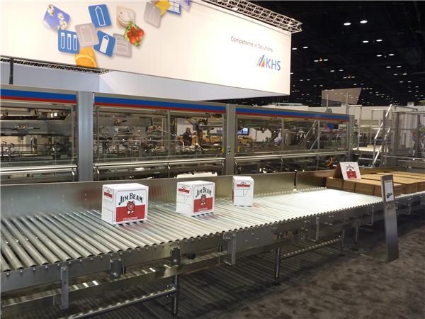 Jim Beam cartons travel along a roller conveyor in front of KHS packaging machinery, under a KHS sign in a large exhibit hall.