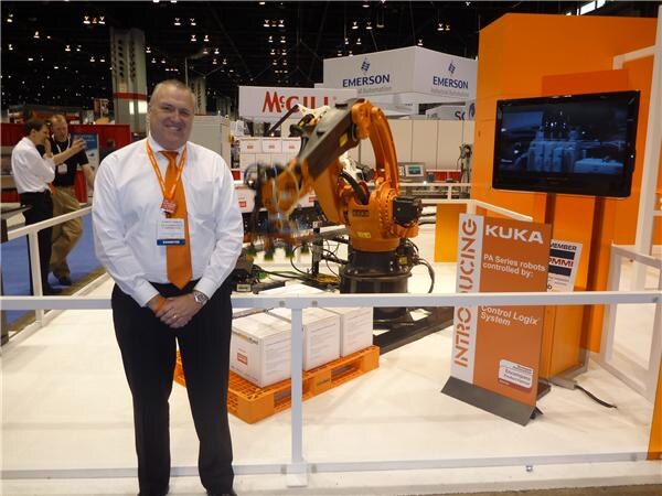 A smiling man in a white shirt and orange tie with a badge stands beside an orange KUKA robot arm and a sign about robots controlled by Control Logix, with Emerson signs behind.