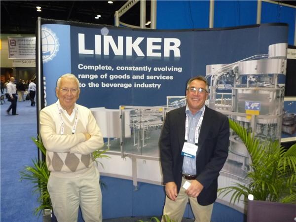 Two men stand in front of a Linker banner reading complete, constantly evolving range of goods and services to the beverage industry. One wears a cream sweater, the other a dark blazer.