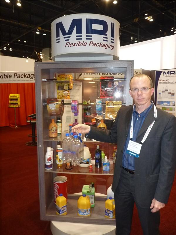A man in a dark jacket and lanyard points to a lit display case of sample bottles, pouches and a Coca-Cola can beneath an MRI Flexible Packaging sign.