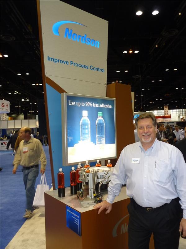 A smiling man in a white shirt stands beside a Nordson display reading Improve Process Control, with a light panel of water bottles and a labeling system showing bottles of orange liquid.